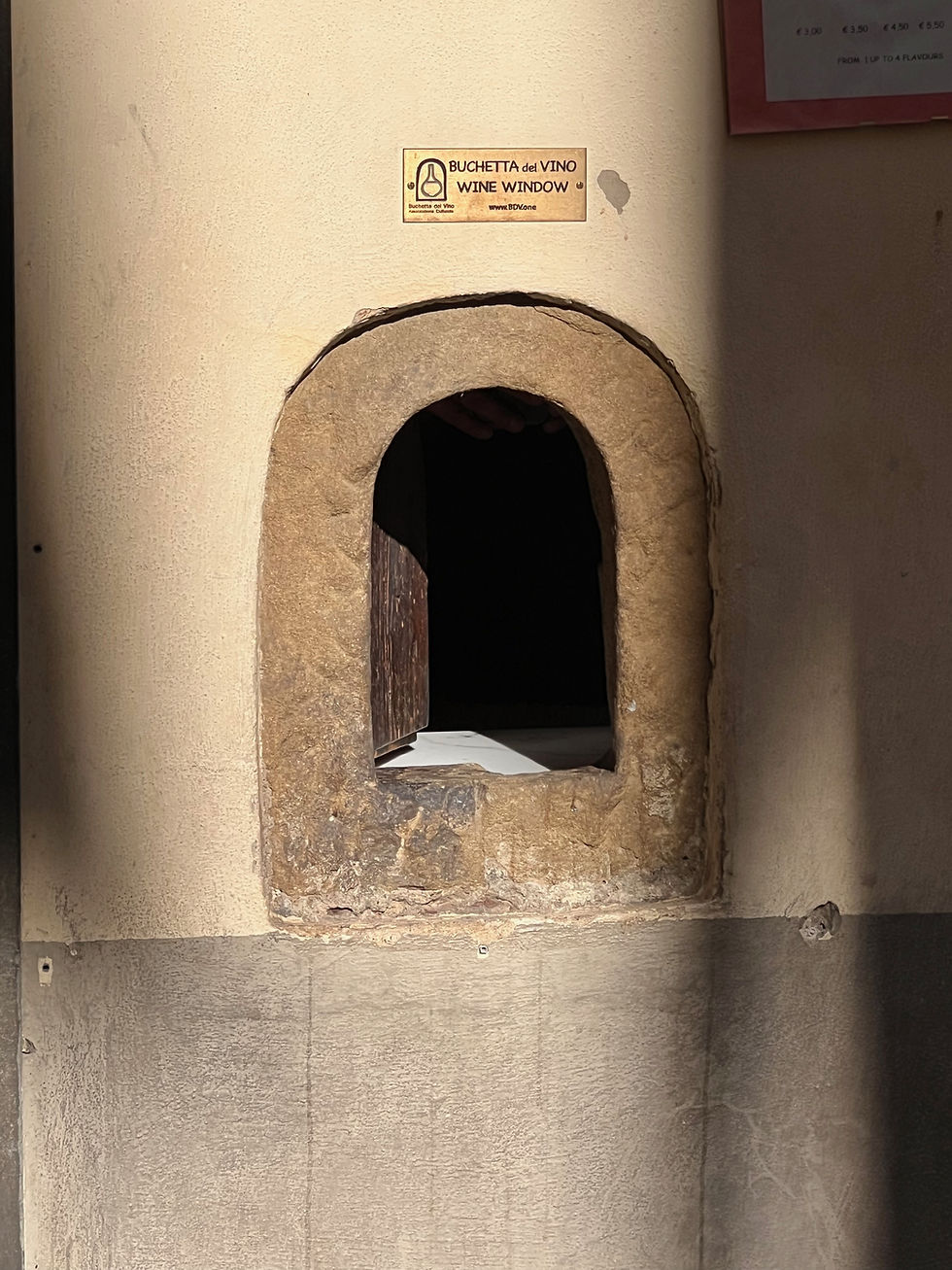 Stone arched wine window in a beige wall with a Buchetta del Vino sign, dark opening, and rustic old-world feel.