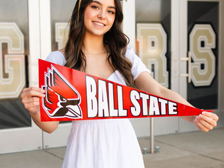 Girl stands in front of Peru High School holding a Ball State University banner in her high school graduation cap.