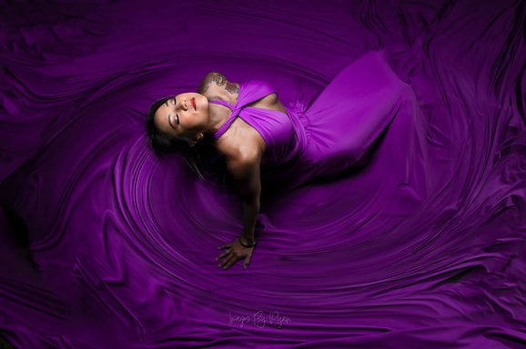 Creative photo session portrait from a high angle, featuring a woman enveloped in a swirling vortex of vibrant purple fabric with dynamic lighting.