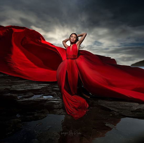 Stunning creative photo session portrait featuring a woman in a dramatic, sweeping red dress against a moody, overcast sky on a rocky shoreline.