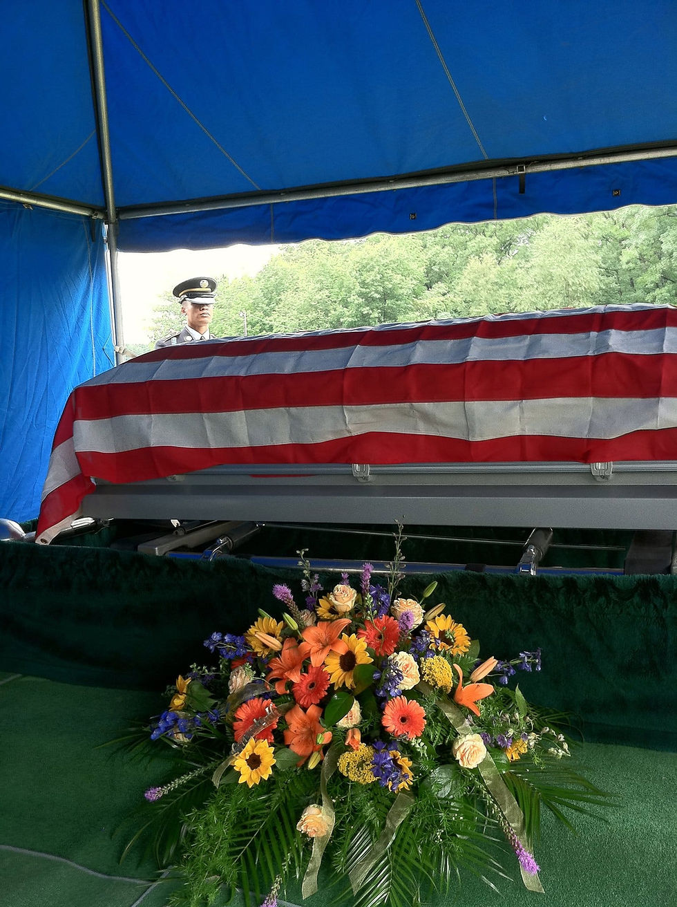 The American flag covered coffin of a WW2 with flowers in front of it is presented with a guard standing in the background.