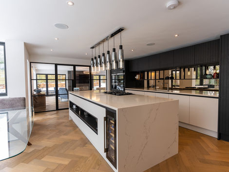 Contemporary open-plan kitchen with large marble-effect island, integrated appliances, wine cooler, and pendant lighting. Features dark wood cabinetry, mirrored splashback, and herringbone wood flooring.