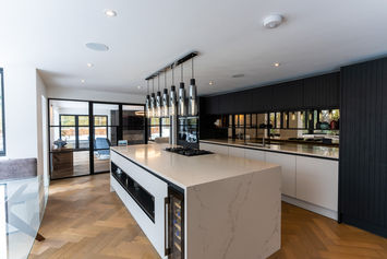 Modern kitchen with suspended glass shelving, white island unit, and dark feature cabinetry enhanced by natural light and parquet flooring.