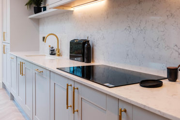 Induction hob area framed by a brushed brass tap, under-cabinet lighting, and pale marble splashback.