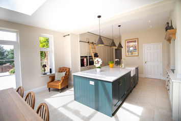 Open-plan kitchen with cream cabinetry, green island, pendant lighting, and oak ladder, designed around an original ceiling beam in Warwickshire home.