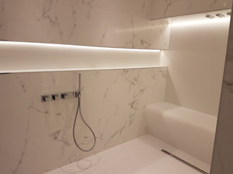 Ceiling-mounted rainfall shower with downlights in a luxurious wet room, surrounded by marble-effect walls and soft ambient lighting.