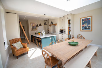 Open-plan kitchen with cream cabinetry, green island, pendant lighting, and oak ladder, designed around an original ceiling beam in Warwickshire home.