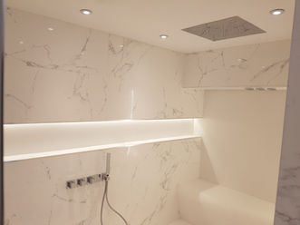 Ceiling-mounted rainfall shower with downlights in a luxurious wet room, surrounded by marble-effect walls and soft ambient lighting.