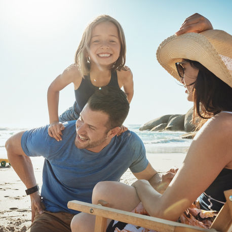 Smiling family at a sunny beach. A child on a man's shoulders, woman in a straw hat, and another child playing with a toy wagon in the background.