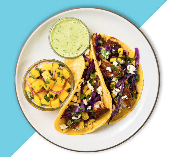 Tacos on a white plate with a bowl of green sauce and yellow fruit cubed