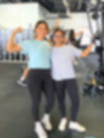 Two women at the gym, smiling. One flexes her arm. They're in activewear with gym equipment and lockers in the background. Energetic mood.