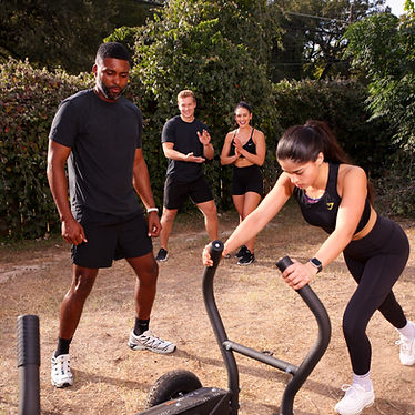 Outdoor workout with a woman pushing a sled while others cheer her on; a man stands clapping in the background.