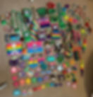 A colorful collection of kandi creations laid out on a carpet, including beaded bracelets, necklaces, cuffs, keychains, and pixel-art designs made from Perler beads. The pieces feature rainbows, skulls, hearts, mushrooms, and other fun rave-inspired symbols in bright neon colors.