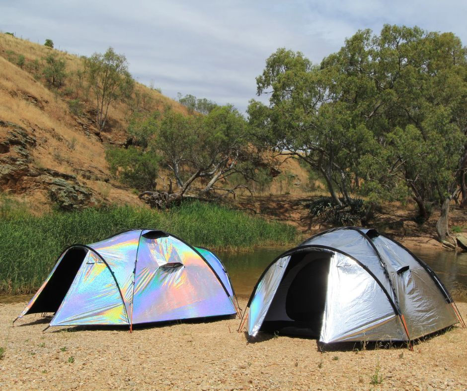 Two Siesta4 festival tents set up in a scenic outdoor location, showcasing one in a holographic finish and the other in a reflective silver design. The tents are surrounded by trees and dry, grassy hills, illustrating their suitability for hot-weather camping.
