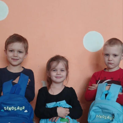 Three smiling children wearing blue dinosaur backpacks against a peach wall.