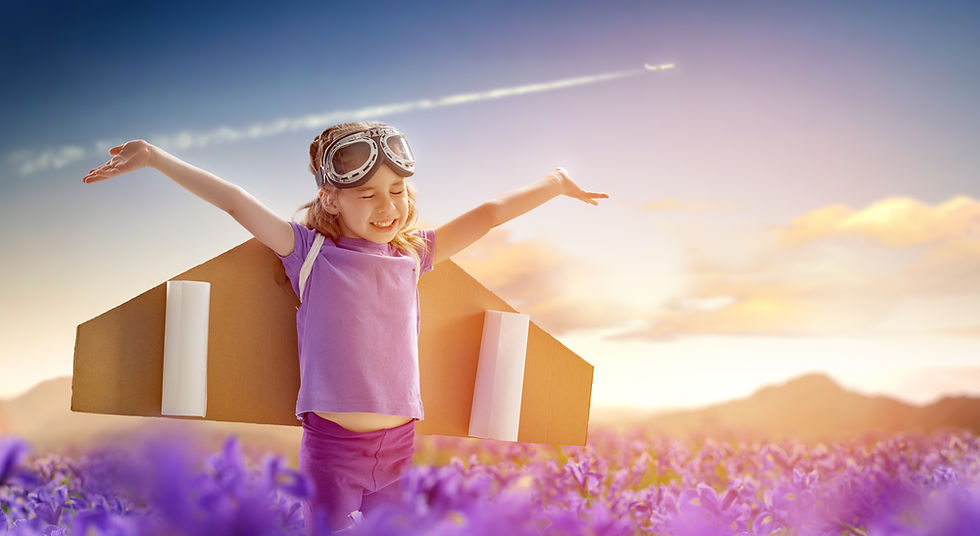 Young girl with outstretched arms wearing cardboard wings, standing in a field of purple flowers under a glowing sky — symbolizing imagination, hope, and quiet strength.