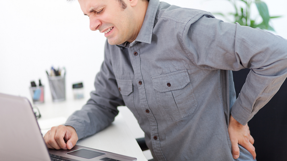 A man sitting at a desk, with his hand on his back, suffering from back pain.