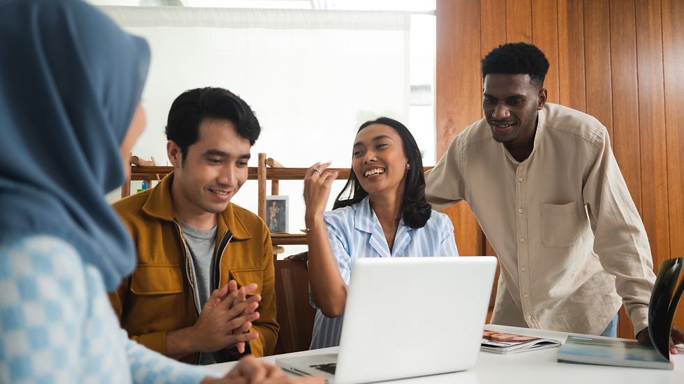 Four people happily discuss around a laptop in a bright room with wooden walls. A woman in a blue blouse laughs, creating a cheerful mood.