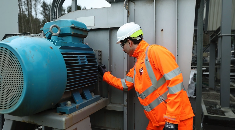 Worker in bright orange safety gear inspects a large blue motor outdoors, surrounded by industrial equipment, wearing helmet and gloves.