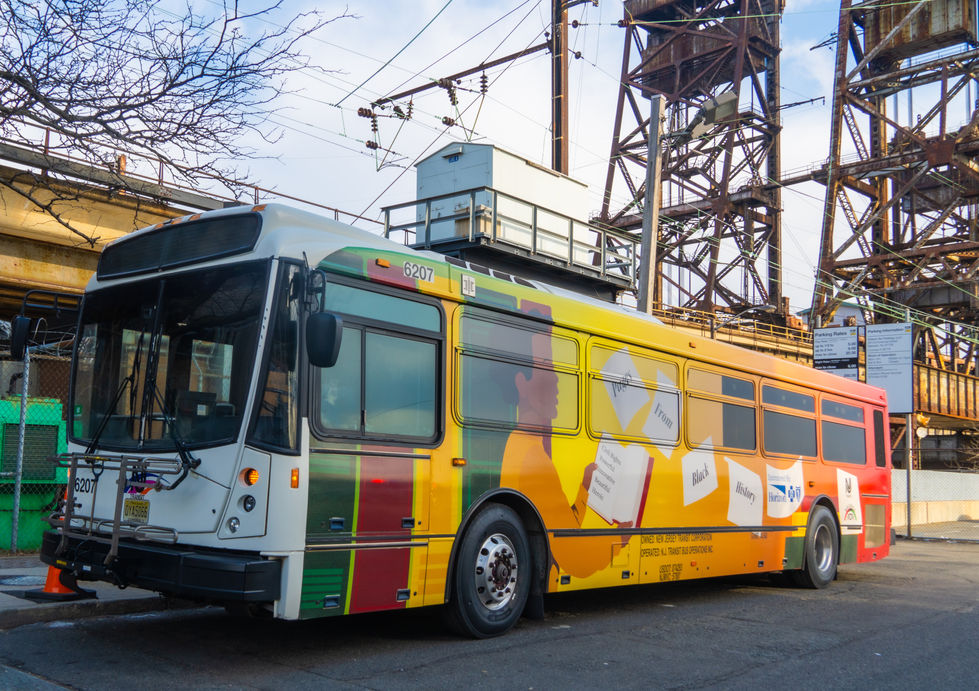 An NJ Transit bus decorated with a Black History Month wrap sits on the street below the train tracks. The bus is covered with colorful book-spine graphics featuring the names of prominent Black historical figures and a large illustration of a child reading. Buildings, utility poles, and rail infrastructure are visible in the background.