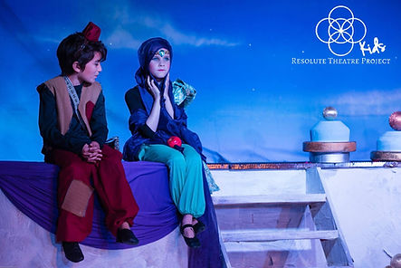 Young kids on stage in colorful costumes acting on stage in Aladdin Jr. musical.