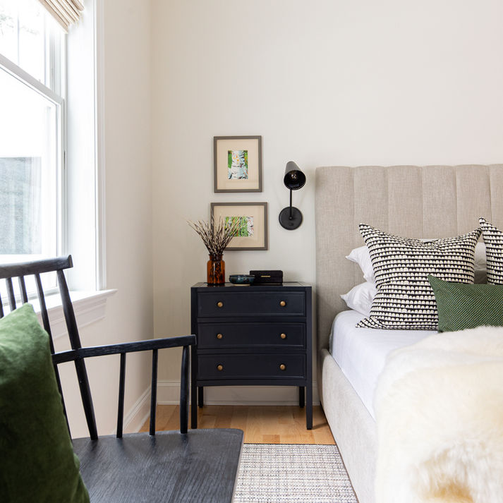 A primary bedroom designed with a beige upholstered bed, a black nightstand with two art prints framed above and a black wall sconce. 