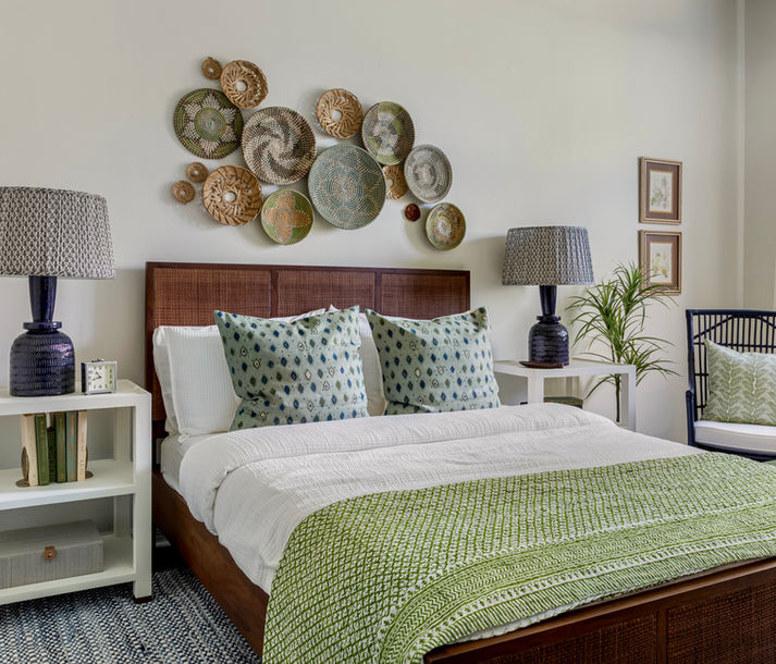 Bright, inviting bedroom with a woven wood bed frame, crisp white bedding, and green patterned pillows and throw. A curated wall installation of woven baskets adds texture above the bed, flanked by matching dark ceramic lamps on white nightstands, with a plant and neutral accents completing the space.