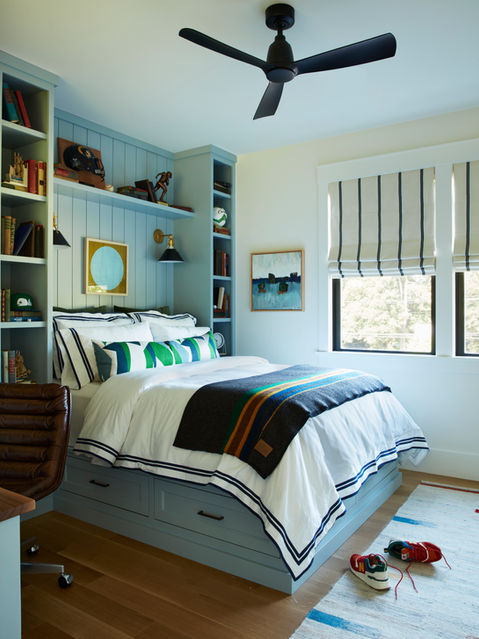 A teenage boys bedroom with a custom built platform bed for lots of storage and two bookshelves on either side housing photo frames, books, mementos and keepsakes. Original art on the window wall is by Kate Pelletier.