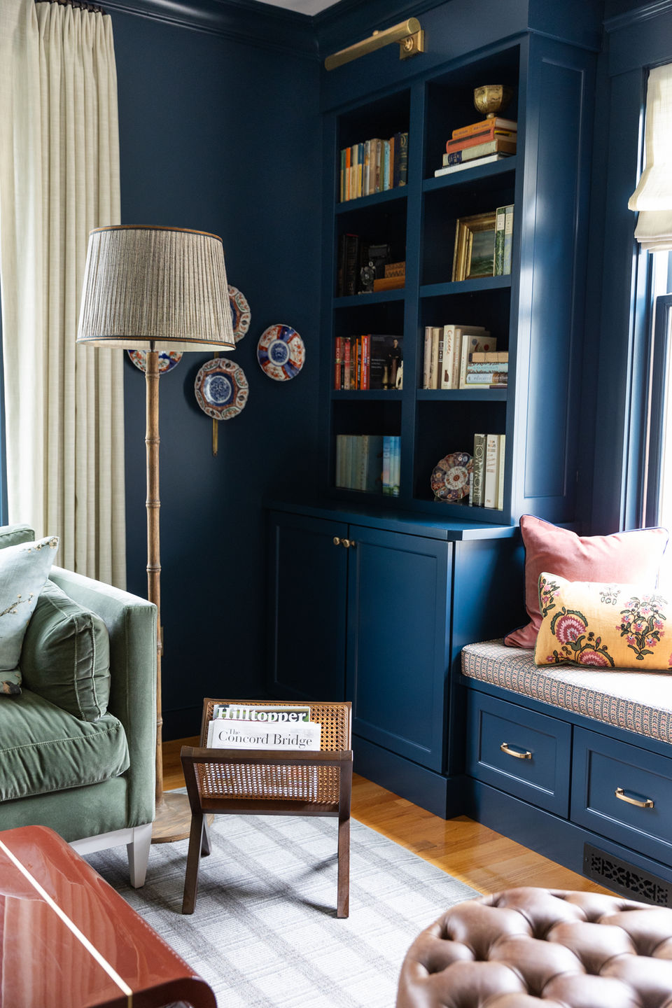 Corner view of a living room with blue built-in bookshelves filled with books and decorative accents, a green sofa, a tall standing lamp, a vintage cane magazine rack and leather ottoman.