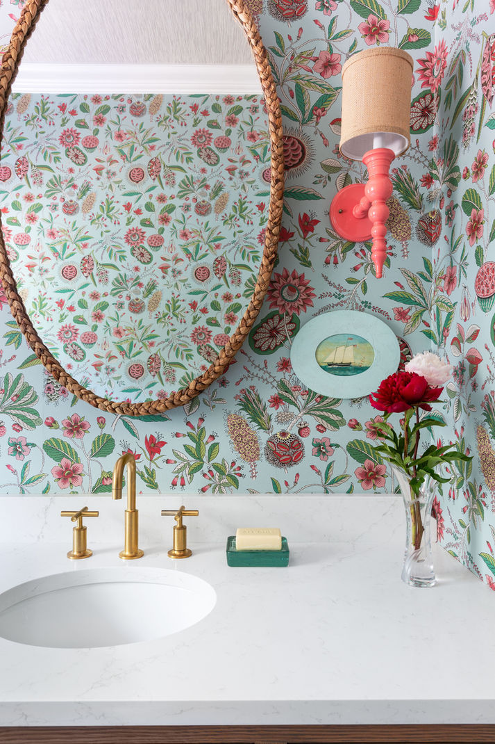 A hall bathroom designed with a floral blue, green and red toned wallpaper, an oval rope mirror and two Dunes and Duchess sconces in coral. 