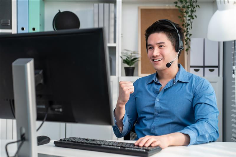 Man in blue shirt wearing a headset, smiling and raising his fist at a desk with a computer in a bright office setting.
