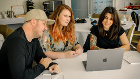 Three SixPivot team members collaborate around a laptop at the SixPivot Summit, engaged in discussion at a white conference table.