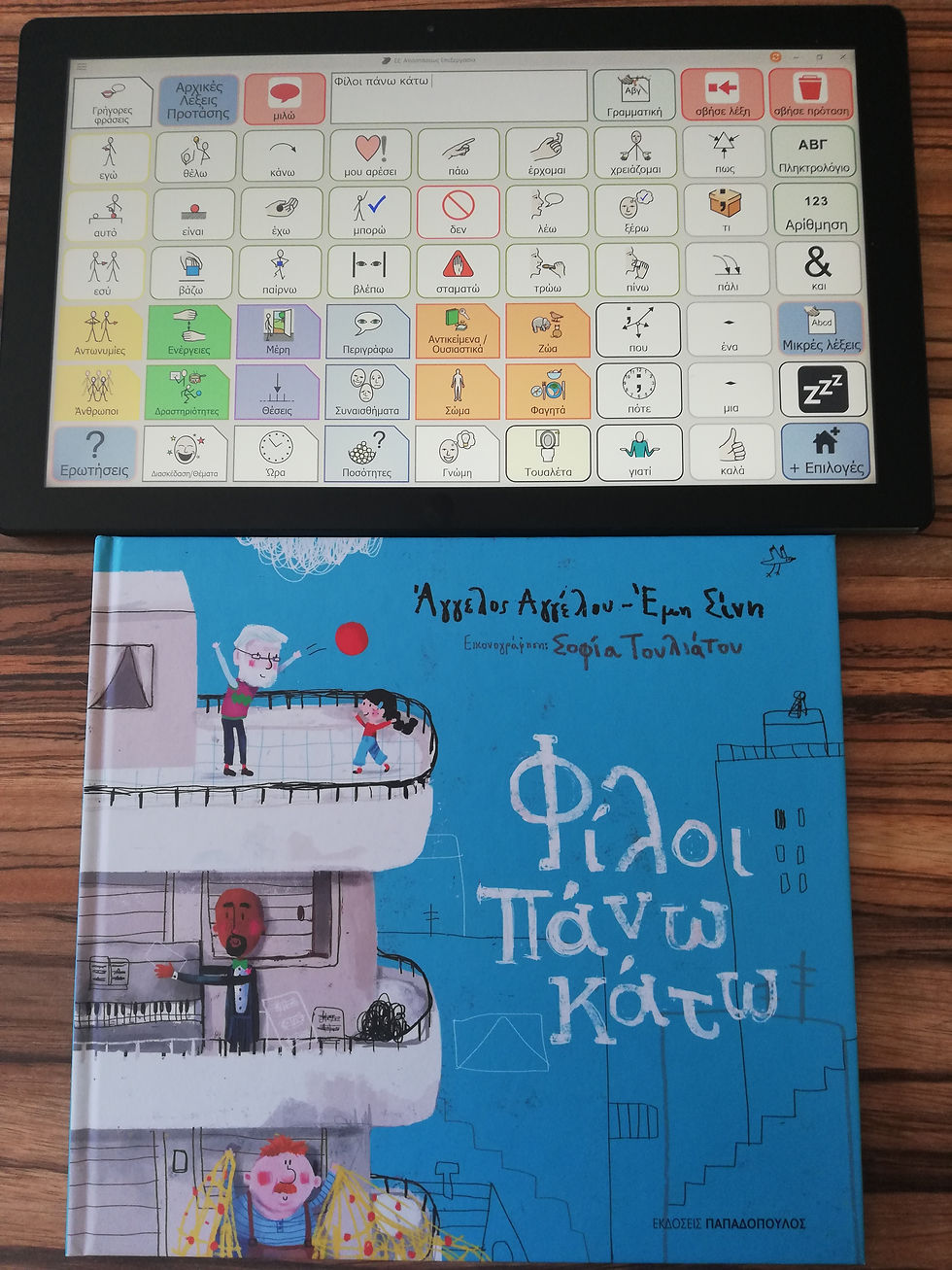Το βιβλίο "Φίλοι πάνω κάτω" και ένα tablet με λεξιλόγιο εναλλακτικής επικοινωνίας πάνω σε ένα τραπέζι. Στο κουτί ομιλίας του γράφει τον τίτλο του βιβλίου.