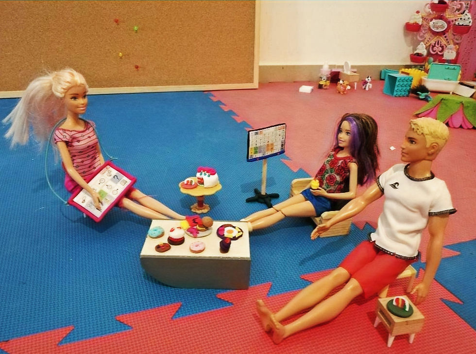 Παιχίδι ρόλων με κούκλες Barbie. Οι κούκλες κάθονται γύρω από ένα τραπέζι με γλυκά, έχοντας μαζί συστήματα εναλλακτικής επικοινωνίας.