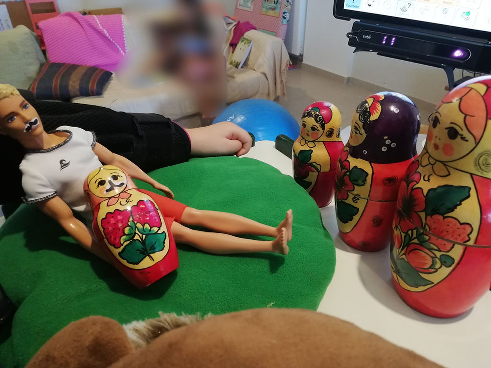 Πάνω στο τραπέζι ενός ορθοστάτη φαίνονται τρεις όρθιες μπάμπουσκες, ένας κούκλος Κεν με αυτοσχέδιο χάρτινο μουστάκι που κάθεται σε ένα μαξιλάρι και έχει στην αγκαλιά του μια μικρή κούκλα μπάμπουσκα με αυτοσχέδιο μουστάκι. Στην άκρη φαίνεται το κάτω μέρος από ένα σύστημα εναλλακτικής επικοινωνίας.