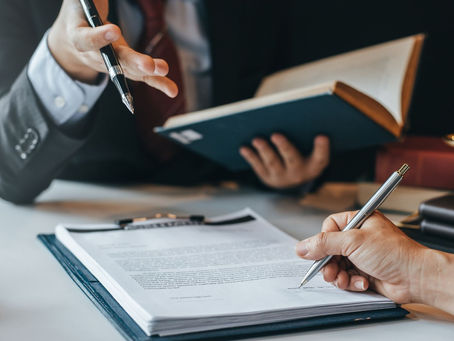 Lawyers in suits review and sign a contract at a desk, with scales of justice and stacked books in a formal office.