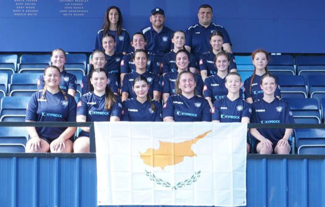 The Cyprus Women's Rugby squad poses for a photo in the stands with the Cyprus flag.