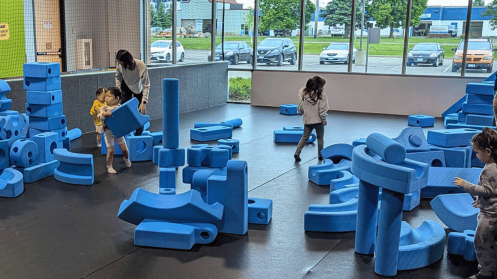 Children and an adult play with large blue foam blocks in a bright indoor play area. Cars and trees visible through large windows.