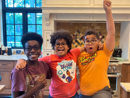 Photo of three smiling boys standing in the kitchen with their arms around one another. They are each wearing fake round black eyeglasses from Harry Potter.