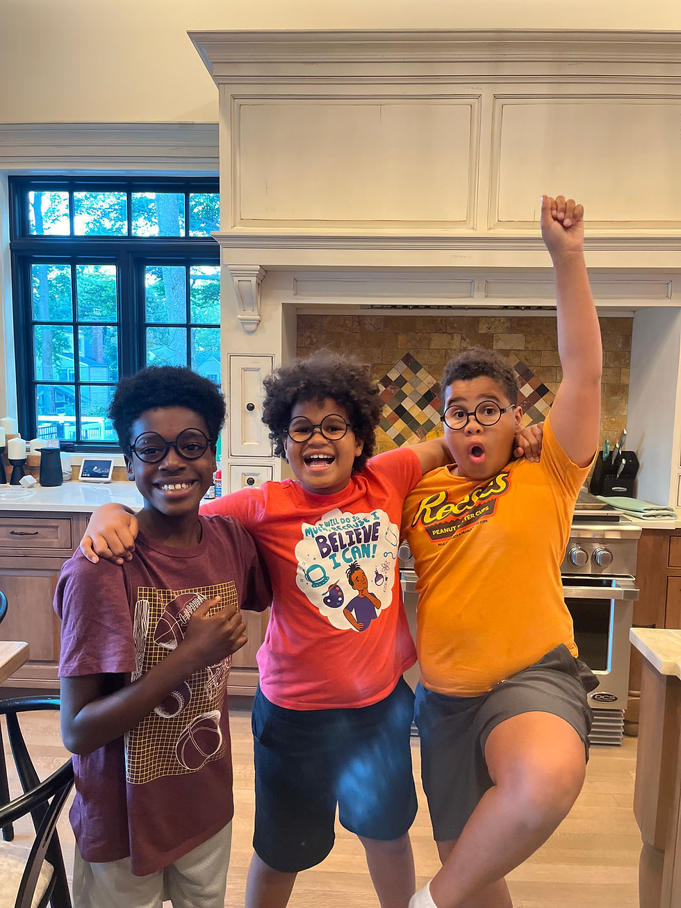 Photo of three smiling boys standing in the kitchen with their arms around one another. They are each wearing fake round black eyeglasses from Harry Potter.