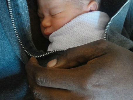 A newborn tucked inside his father's sweatshirt. The baby is cradled by his father's hand.