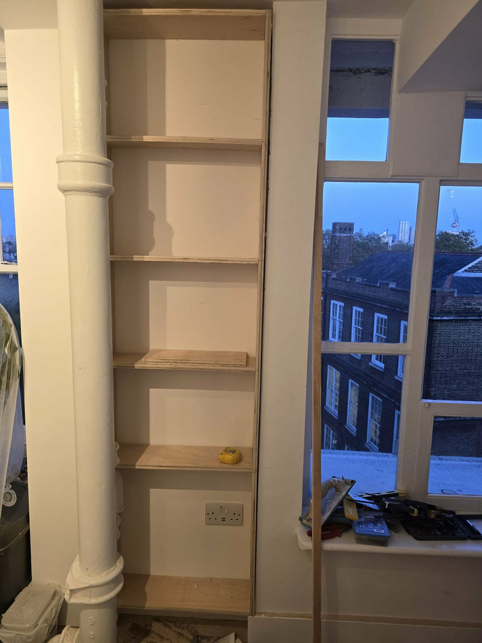 Part‑built shelving unit fitted into a narrow alcove beside existing pipework, showing custom construction to maximise space.