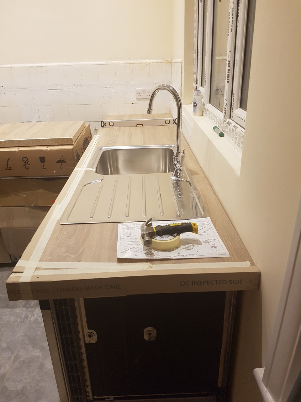 Kitchen installation in progress with a wooden worktop, fitted sink, tools on the surface, and partially tiled backsplash.