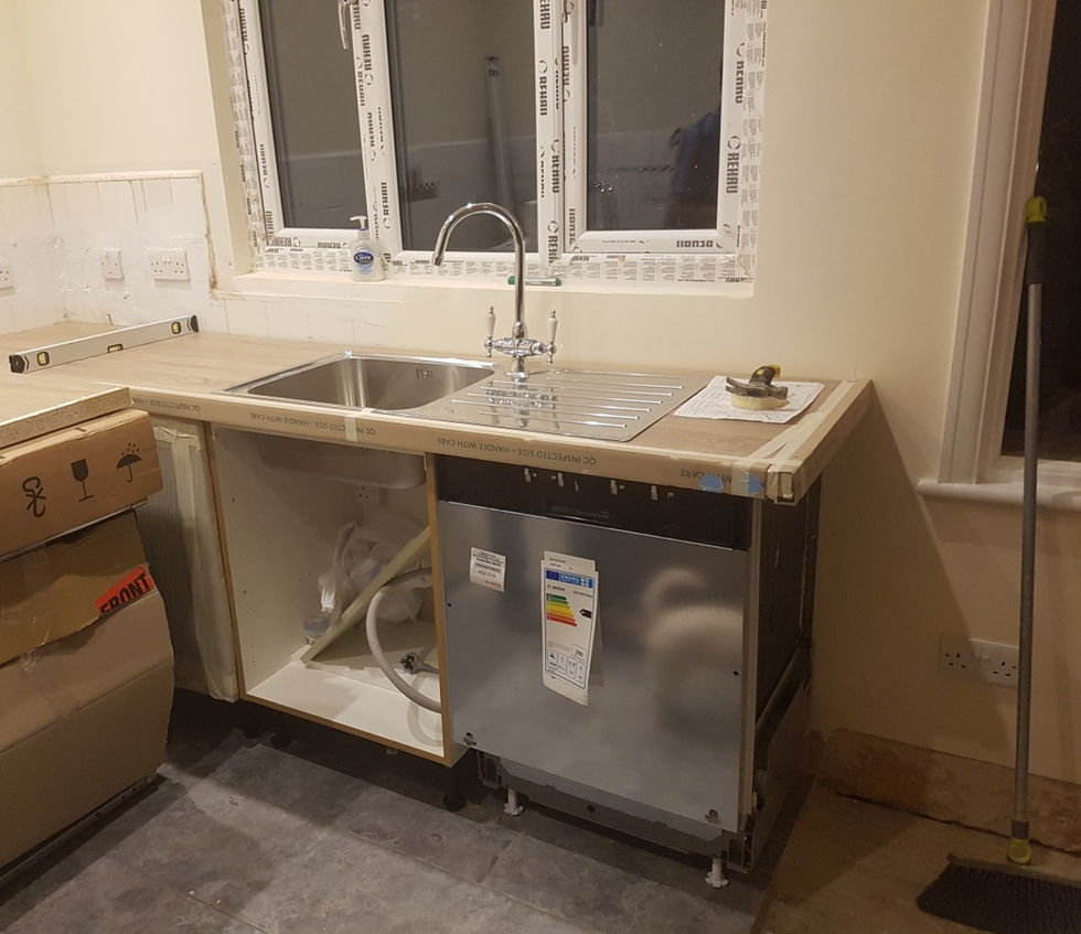 Partially installed kitchen with a new worktop, stainless steel sink, exposed plumbing, and a dishwasher being fitted.