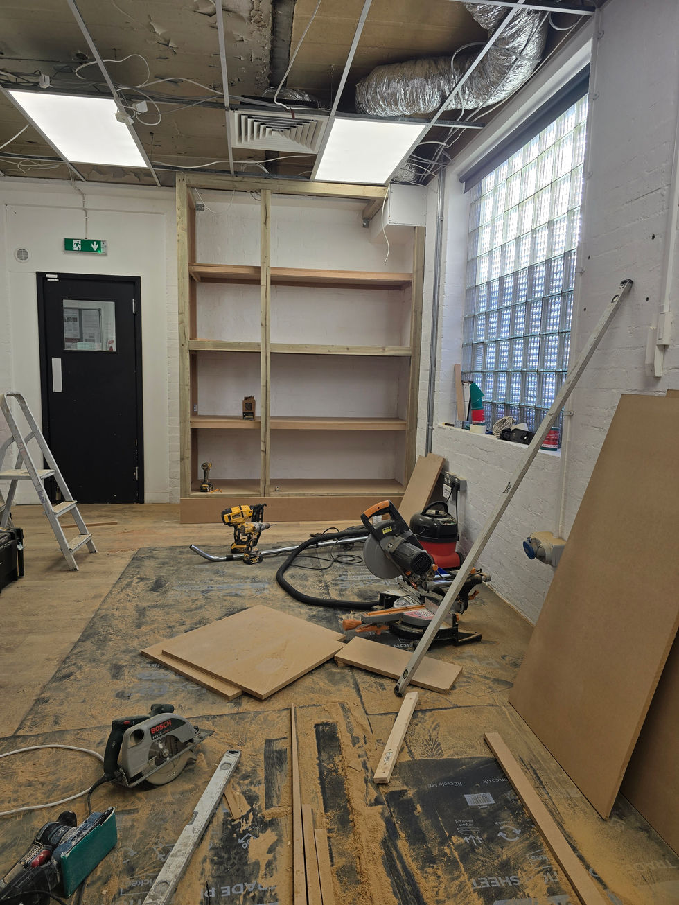 Workshop space with woodworking tools, timber boards, and a partially assembled shelving frame on the floor, showing the early construction stage of the built‑in office shelving and cupboard project.