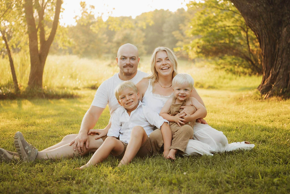 A smiling family. Candidate Shane Sawyer and wife sitting with their two boys in the grass. Warm toned images, family is wearing white and tan.
