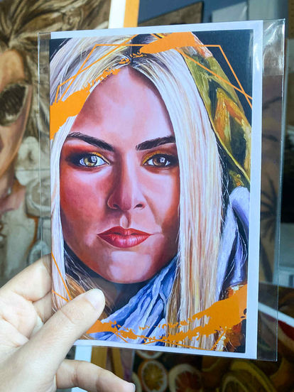 Main tenant une carte de souhait imprimée (sous plastique) avec reproduction d’un portrait artistique aux accents orange, en atelier.