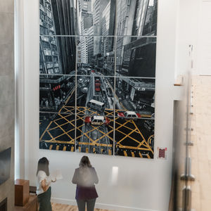 Peinture d’une mosaïque de 12 toiles à l'huile d''un carrefour d'une grande ville en Asie vu du dessus, avec immeubles hauts, circulation et quadrillage jaune au sol, accents rouges sur des bus.