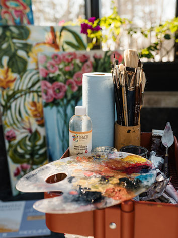 Palette de peinture avec couleurs et pinceaux sur un chariot d’atelier, avec toiles florales et fenêtre en arrière-plan.