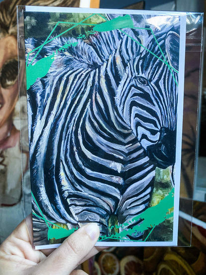 Main tenant une carte de souhait imprimée (sous plastique) illustrant un zèbre en gros plan, style peinture, avec encadrement graphique vert.